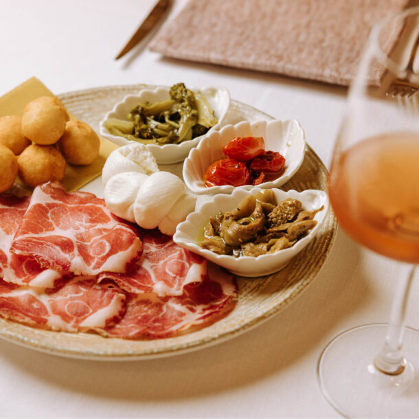 Antipasto pugliese con capocollo, treccia di mozzarella, frittelle e ciotoline di verdure sott’olio, accanto a un calice di rosato